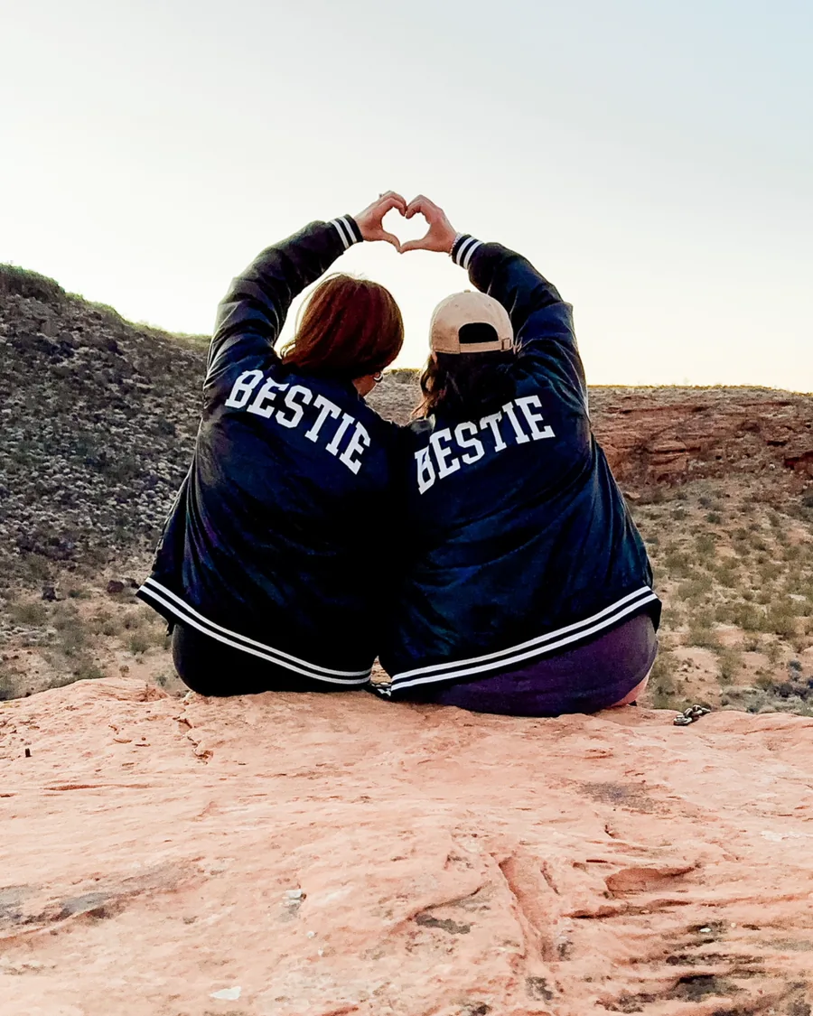 Two Bestie Squad members in BESTIE jackets making a heart with their hands on red rock