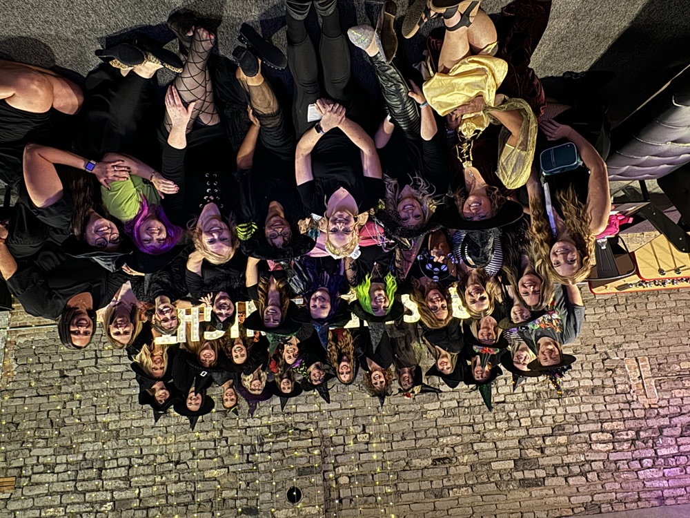 Bestie Squad members in costume at a themed event, packed together against a brick wall for a group photo.